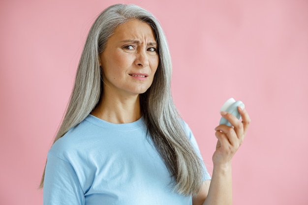 Disgusting grey haired Asian woman holds sonic facial cleansing brush on pink background in studio. Mature beauty lifestyle