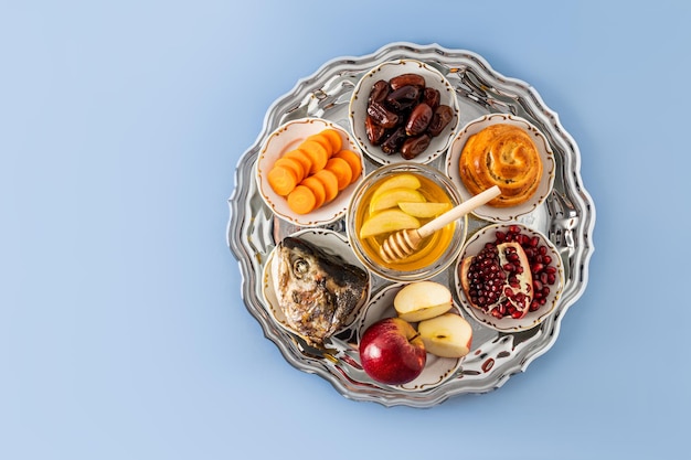 Photo a dish with a new year's treat for the new year's holiday rosh hashanah pomegranate honey dates apple and carrots blue background copy space