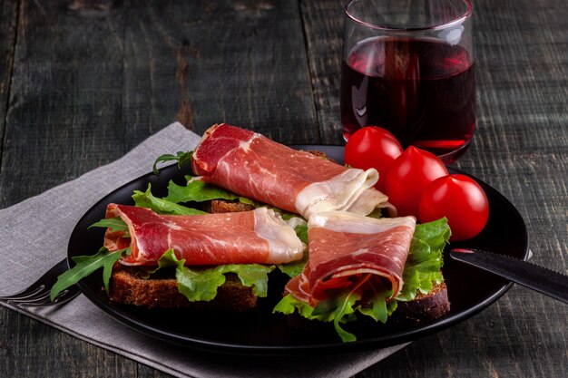 A dish with sandwiches with jamon, herbs, tomatoes and a glass of wine stand on a wooden table