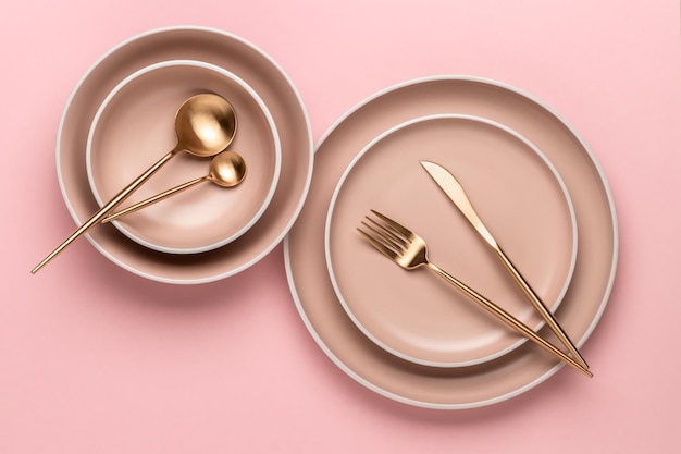 Dishes and utensils for serving and eating meals Beige round rimmed plates and golden cutlery on a pink background top view Modern craft ceramic crockery trendy tableware from above