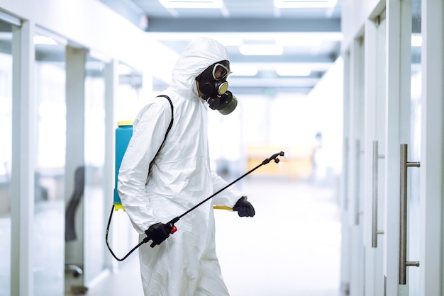 COVID-19を防ぐためのオフィスの消毒、スプレー化学物質で保護化学防護服を着た男。