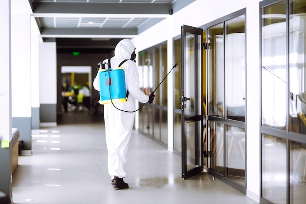 Дезинфекция офиса для предотвращения COVID19 Человек в защитном костюме химзащиты с распыляющими химикатами