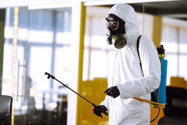 Дезинфекция офиса для предотвращения COVID19 Человек в защитном костюме химзащиты с распыляющими химикатами