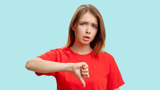 Dislike gesture negative answer portrait of annoyed skeptic\
woman in red tshirt criticizing showing thumb down isolated on blue\
background loser shame mistake failure blame