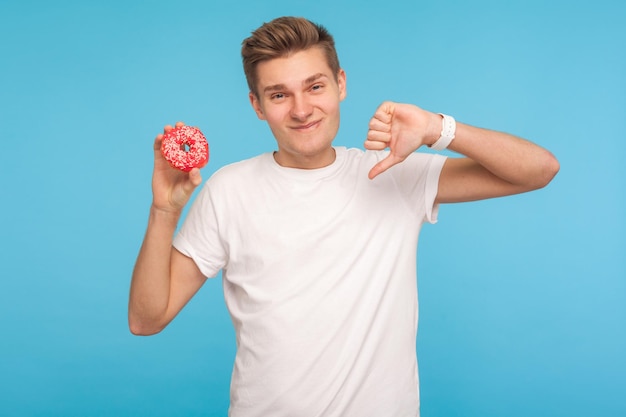 ジャンクフードが嫌いTシャツを着た不満な不幸な男が親指を下に向けて甘いドーナツを持って高カロリーのデザートについて警告している不健康な栄養屋内スタジオショットブルーの背景