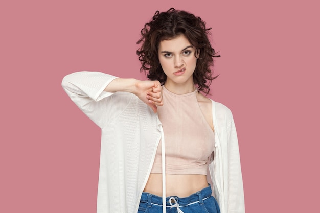 Dislike. Portrait of dissatisfied beautiful brunette young woman with curly hairstyle in casual style standing, looking at camera, holding thumbs down. indoor studio shot isolated on pink background.