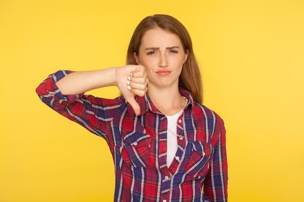 Dislike Portrait of dissatisfied unhappy ginger girl in red shirt showing thumb down gesture and looking displeased negative feedback disapproval sign studio shot isolated on yellow background