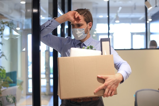 Foto licenziamento dipendente in maschera medica preventiva in un coronavirus epidemico. lavoratore licenziato che va dall'ufficio con le sue forniture per ufficio.