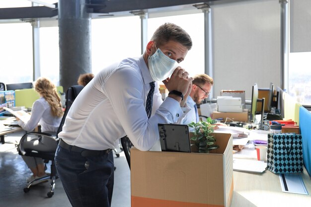 Foto licenziamento dipendente in maschera medica preventiva in un coronavirus epidemico. il lavoratore licenziato triste sta portando con sé le sue forniture per ufficio dall'ufficio.