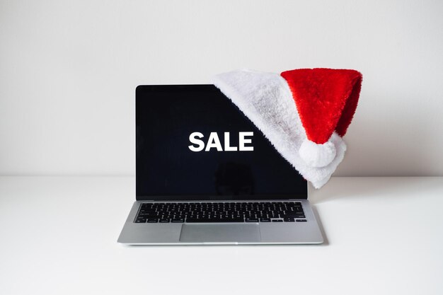 Экран ноутбука с распродажей слов и шляпой санта-клауса на белом столе праздничный домашний офис