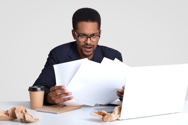 Displeased African American man looks with puzzlement at paper documents, has deadline to prepare financial report, sits at destop agaist white wall, works on laptop computer. Paperwork