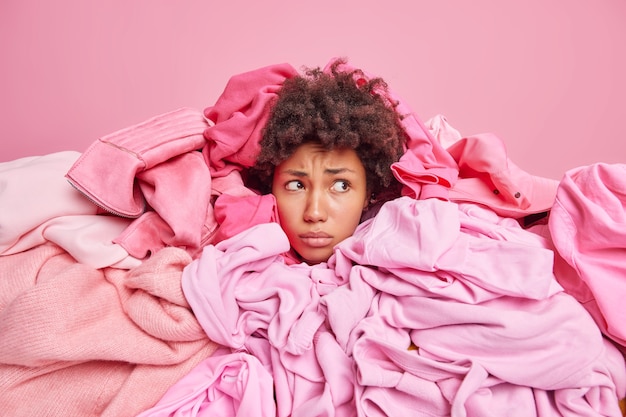 Displeased African American woman focused away with unhappy expression covered with big heap of clothes busy doing laundry isolated over pink wall. Stack of clothing thrown on female model