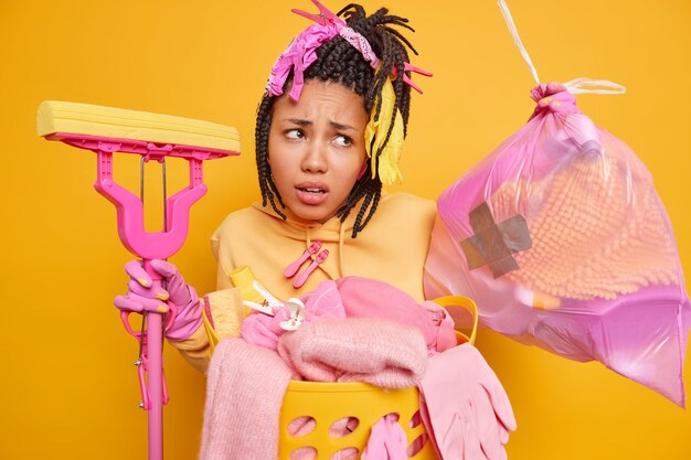 Photo displeased african american woman with dreadlocks busy doing housework holds mop and garbag