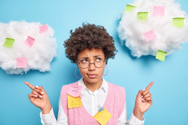 Displeased Afro American woman points sideways with clueless expression shares ideas on colorful sticky notes wears round spectacles poses against blue wall