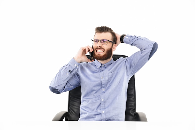 Displeased bearded elegant man in eyeglasses talking by smartphone while sitting by the table