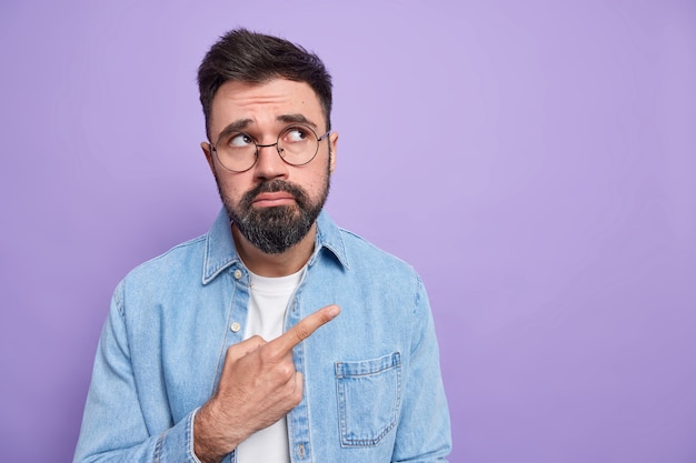 Displeased bearded man purses lips doesnt like something has sulking disappointed face expression wears spectacles denim shirt 