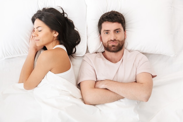 a displeased confused young man lies in bed under blanket near sleeping woman