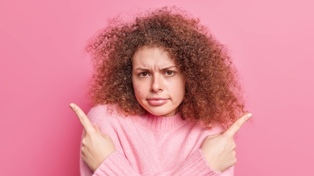 Displeased curly haired puzzled young woman points right and left hesitates between two options has grumpy face expression dressed casually isolated over pink wall. What should I choose