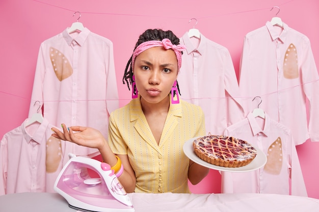 Displeased dark skinned young woman looks puzzled at camera raises palm bakes delicious pie irons laundry at home does domestic chores. Busy housewife has many tasks to do