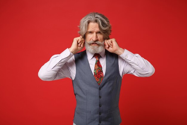 Displeased elderly gray-haired mustache bearded man in classic shirt vest colorful tie isolated on red background in studio. People lifestyle concept. Mock up copy space. Covering ears with fingers.