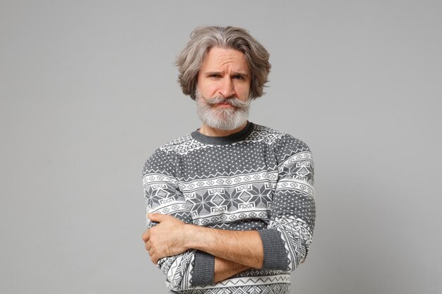 Displeased elderly gray-haired mustache bearded man in knitted\
sweater posing isolated on grey wall background, studio portrait.\
people lifestyle concept. mock up copy space. holding hands\
crossed.