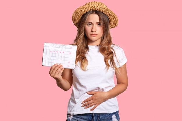 Photo displeased european young female feels pain and spasm during menstruation period, holds calendar, keeps hands on bottom of belly, wears straw hat and white t shirt, isolated over pink wall.
