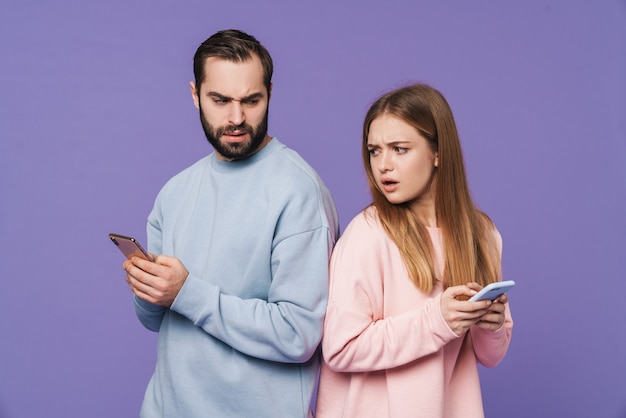 displeased girl looking at her man chatting by mobile phone isolated over purple wall.