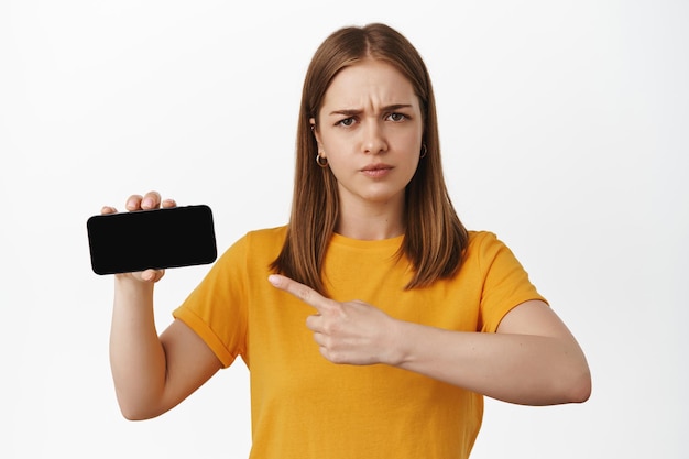 Photo displeased girl pointing at smartphone screen, frowning upset, asking question about smth strange online, showing promo on phone, standing against white background