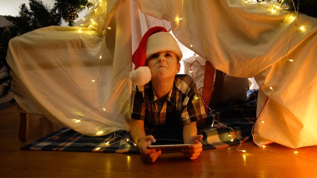 Displeased little boy in Santa hat lying in tent using phone Christmas concept