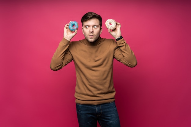 Displeased man holds two tasty ring donuts, feels unhappy as cant eat sweets, isolated over pink background.