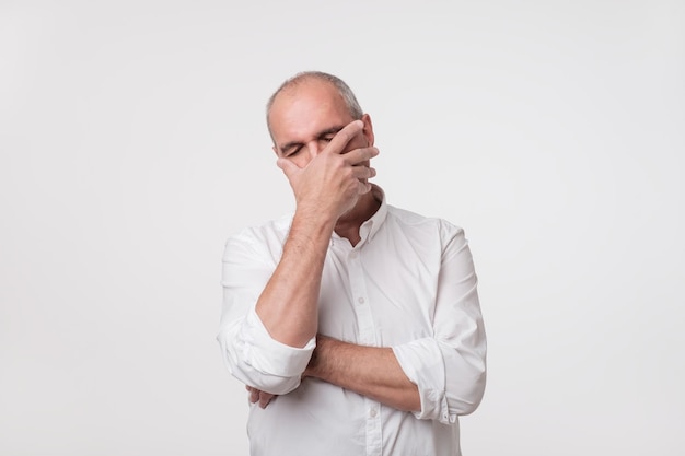 Displeased mature man in white shirt covering his face with hand over gray background