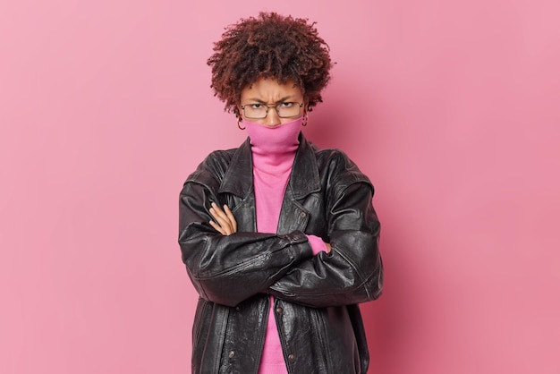 Displeased offended woman crosses arms and looks angrily at camera has grumpy discontent expression covers mouth with collar of jumper wears spectacles and black leather jacket isolated on pink wall