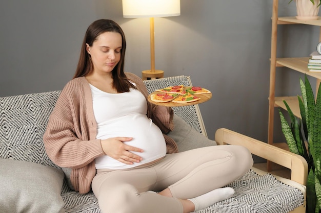 Photo displeased pregnant woman eating pizza alone at home sitting on sofa in living room overeating fast food during pregnancy touching her belly feeling discomfort