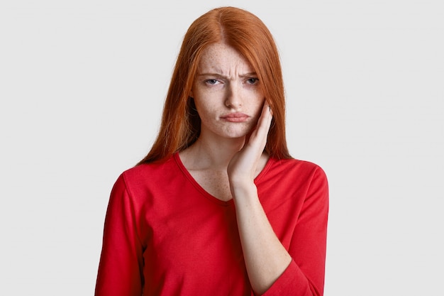 Displeased red haired woman with freckled skin, keeps hand on cheek, suffers from toothache, has sensitivity, wears casual red clothes, isolated on white. Dental problems concept