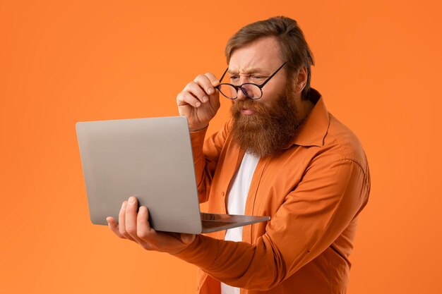 Displeased redhaired bearded man looking at laptop through eyeglasses studio