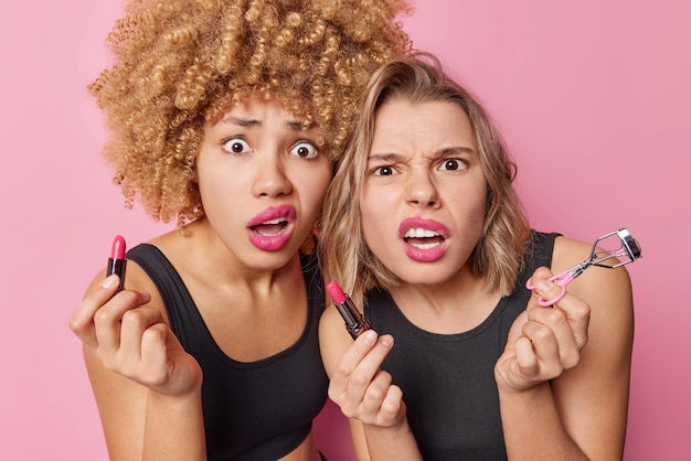 Foto le donne scioccate dispiaciute applicano il rossetto rosato si truccano prima dell'occasione speciale usano il bigodino delle ciglia indossano magliette nere isolate su sfondo rosa si fissano allo specchio essendo a corto di tempo