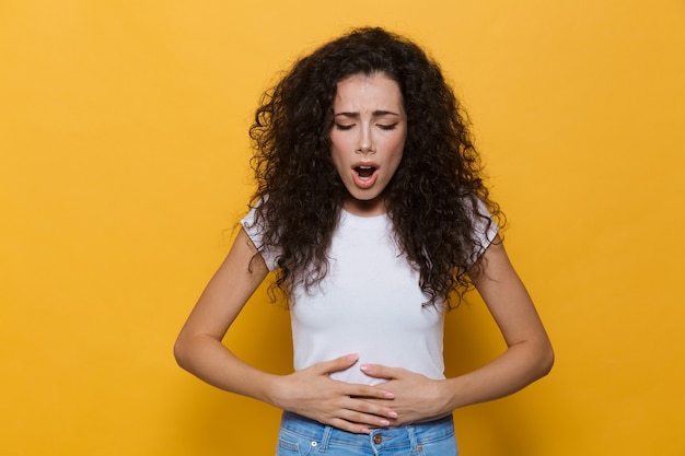 displeased woman 20s with curly hair touching stomach and bending isolated on yellow