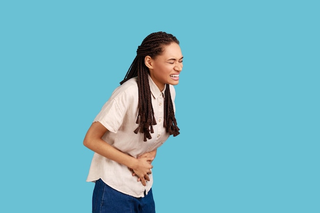 Displeased woman with black dreadlocks feels discomfort in stomach, keeps palms on tummy, has period cramps, ate spoiled food, wearing white shirt. Indoor studio shot isolated on blue background.