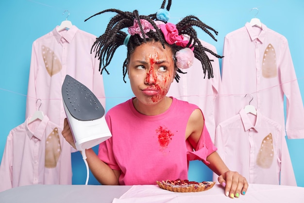 Displeased woman with braided hairstyle has jam on face after\
eating pie holds electric iron looks sadly aside stands near\
ironing board in laundry room dreams about rest after doing all\
housework