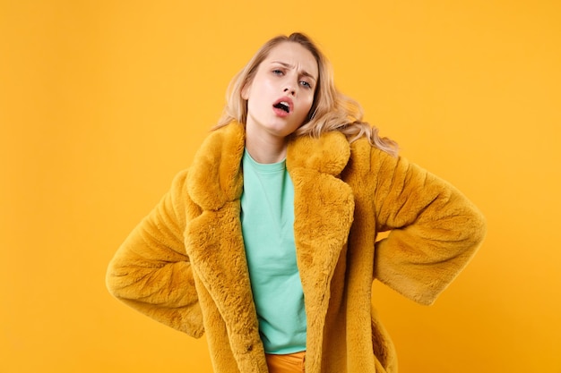 Displeased young blonde woman girl in yellow fur coat. posing isolated on orange background studio portrait. people sincere emotions lifestyle concept. mock up copy space. standing with arms akimbo