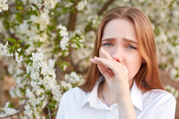 La giovane donna dispiaciuta ha il naso che cola, si sente allergica ai fiori di stagione durante la primavera, vestita con una camicia bianca, non posso camminare all'aperto