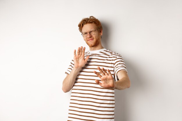 Displeased young man with red hair and bristle showing stop gesture, refusing something bad with disgusted face, standing over white background.