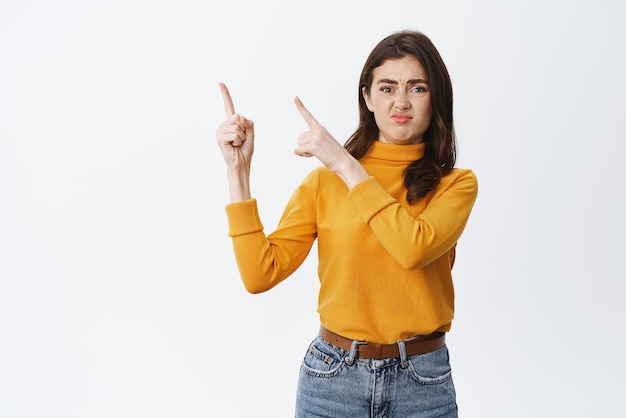 Displeased young woman client complaining pointing at upper left corner with aversion and dislike on face show bad thing standing on white background