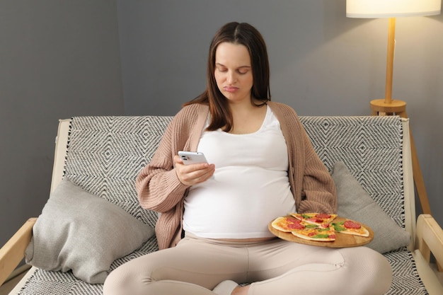 Недовольная молодая женщина, сидящая на диване с тарелкой с пиццей, использующая смартфон, просматривающая веб-страницы Интернета, в то время как ест нездоровые закуски, используя мобильный телефон, проверяя скучные социальные сети