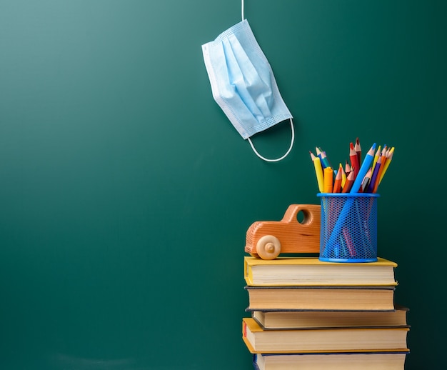 Disposable medical mask, stack of books and school stationery on green chalk board