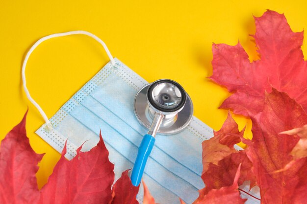 Disposable protective medical mask and stethoscope and autumn maple leaves on yellow background