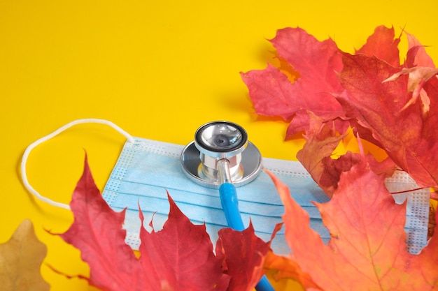 Disposable protective medical mask and stethoscope and autumn maple leaves on yellow background