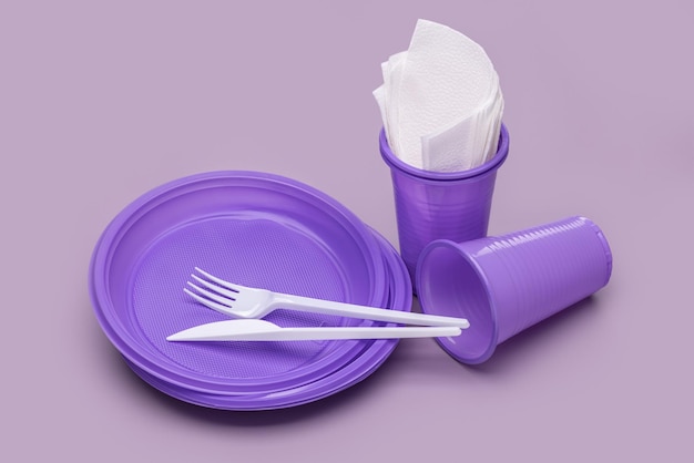 Disposable purple plates, glasses for drinks and cutlery on a\
pink background, paper napkins. studio