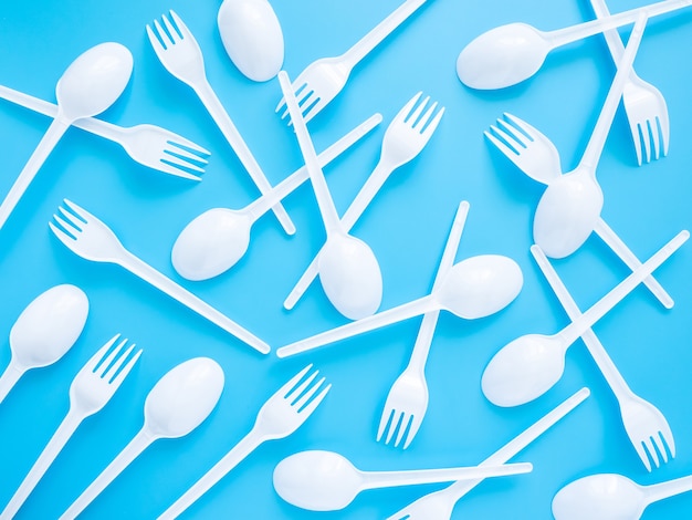 Disposable tableware, white plastic forks and spoons are scattered on a blue background, top view, flat lay.