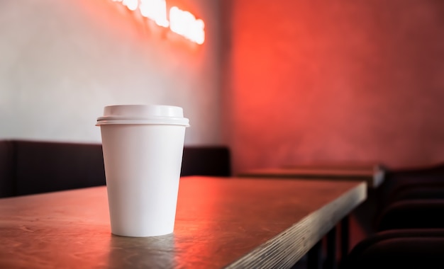 Disposable white paper coffee cup mock up stands on the table with neon light.